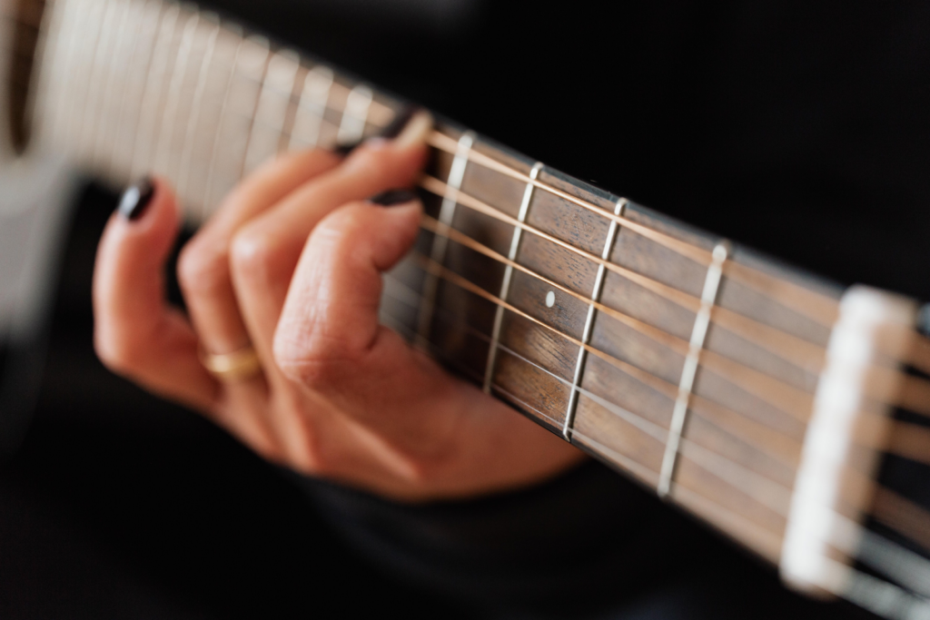 A close up of the fretboard of a guitar, with a woman with black nail polish holding the guitar.