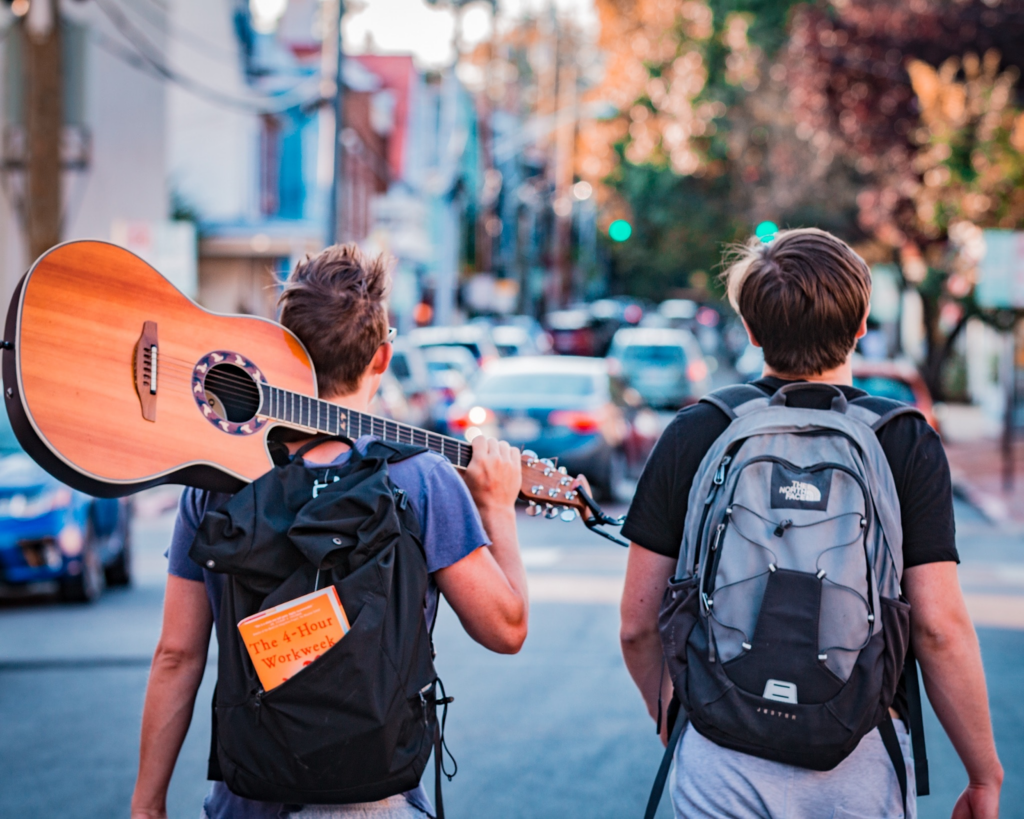 Two backpackers walking down a busy road, one of the backpackers is carrying an acoustic guitar over his shoulder.