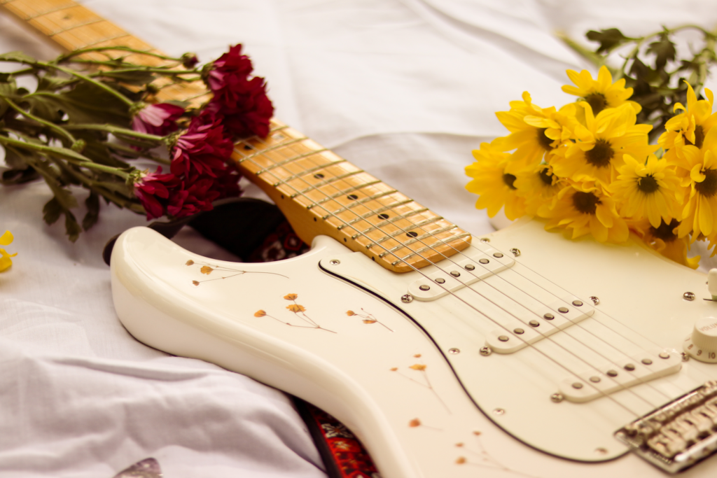 A white electric guitar with a yellow flower print lying amongst bunches of flowers.