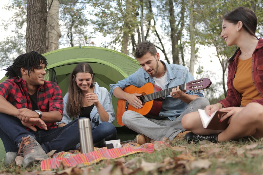 A group of young people at a campsite, listening to a man play guitar.