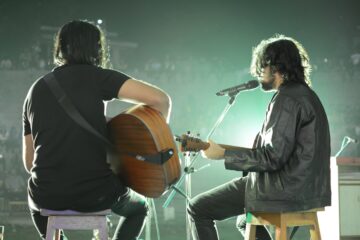 Two men sitting on guitar stools performing on stage.