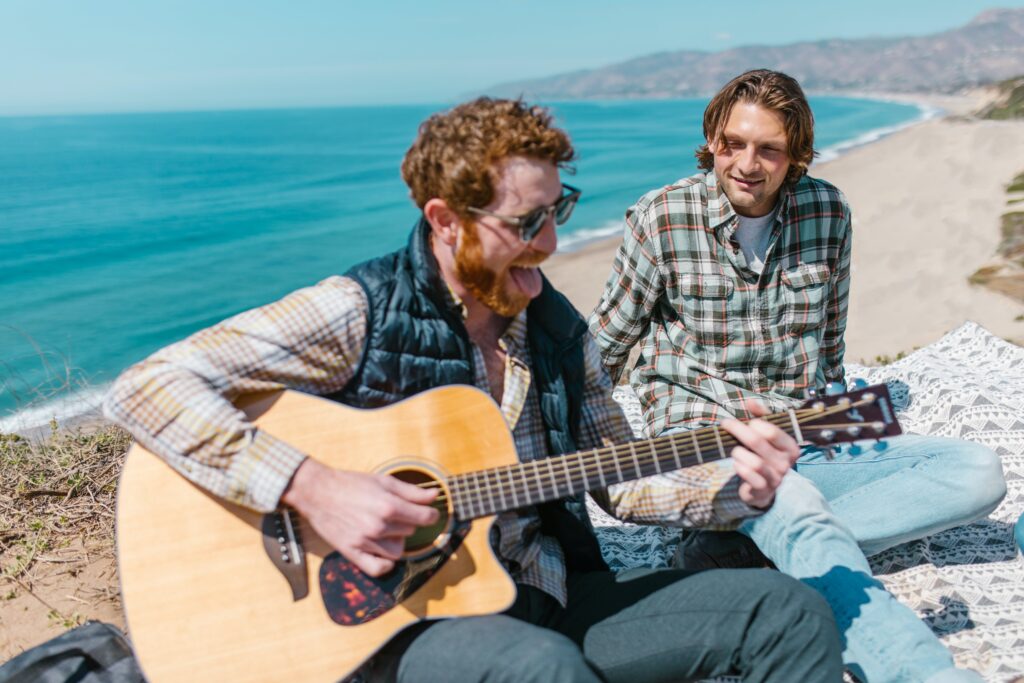Two young men sitting by the seaside, while one man plays the guitar.