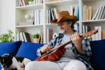 A man in a cowboy hat playing guitar on his couch while his dog listens.