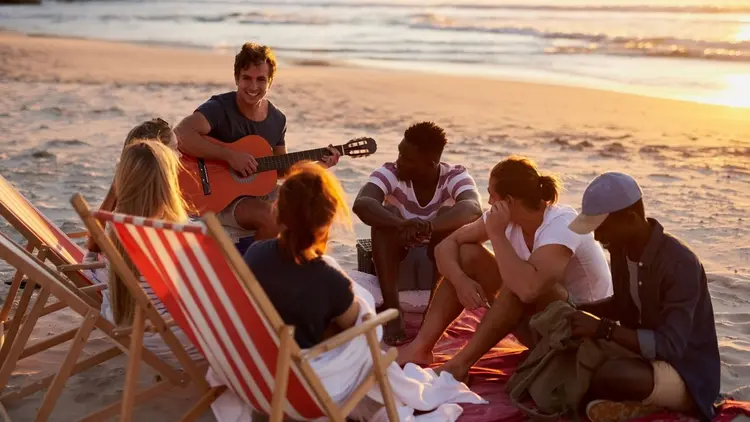 A group of friends at the sea, gathered around a guitar to sing songs.